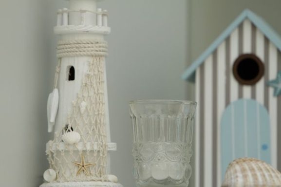 Leuchtturm mit Muscheln, daneben ein Glas und ein blau-weiß gestreiftes Haus.