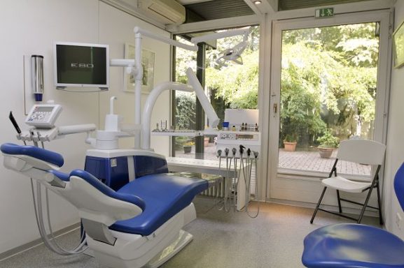 Zahnarztpraxis mit blauen Behandlungsstühlen und Fensterblick auf den Garten.