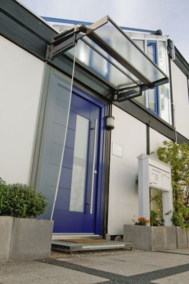 Moderne Haustür mit blauer Farbe und Glasvordach, umgeben von einige Pflanzen.